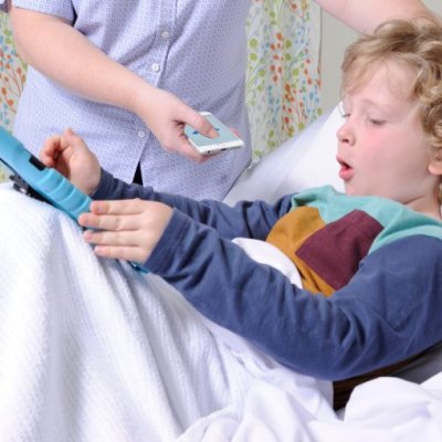 A small boy lies in a hospital bed, while a nurse holds a mobile phone near him to record his cough.